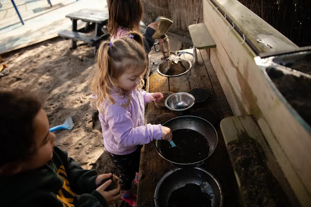 Young children playing wooden sink bench sandpit in background